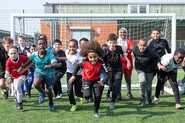 A group of young people of all ages wearing sports clothing, running towards the camera. There is a football goal post in the background.