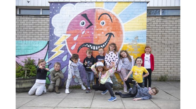 A group of kids posing for a picture The background is a colourful mural which depicts different types of weather. 